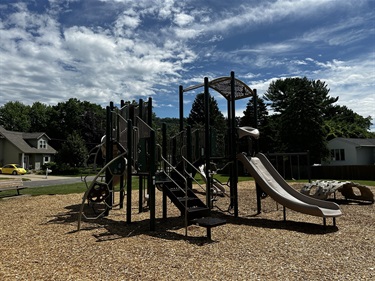 A modern playground with slides, climbing structures, and a tunnel, set on a wood chip ground cover with a blue sky and white clouds above.