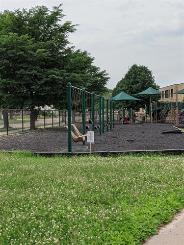 A playground with swings, slides, and climbing structures on a black rubber surface, bordered by green grass and trees.