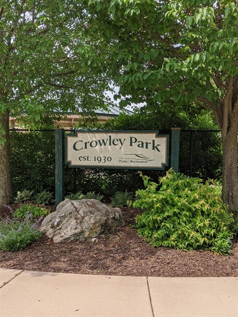 A green and tan playground with slides, swings, and climbing structures sits on dark mulch under a cloudy sky.