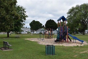 A colorful playground structure with slides and climbing bars sits on a wood chip base in a grassy park. Two benches and a trash can are also visible.