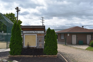 A sign for Erickson Park stands next to a baseball field with a small brown building behind it.