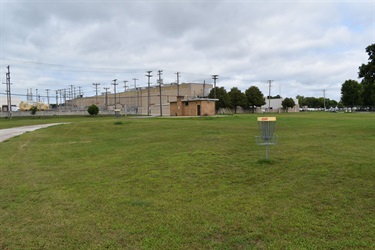 A disc golf basket stands in a grassy field with a large industrial building and power lines in the background under a cloudy sky.