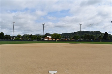 A baseball field with a dirt infield and grass outfield, surrounded by trees and hills under a cloudy sky. Lights for night games are visible along the outfield fence.