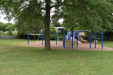 A blue and tan playground with slides, monkey bars, and a climbing net on a sandy surface surrounded by grass and trees.