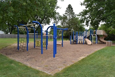 A blue and tan playground with slides, monkey bars, and a climbing net on a sandy surface surrounded by grass and trees.