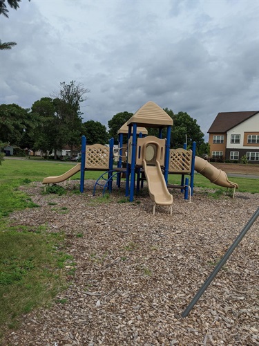 A tan and blue playground structure with two slides, a tunnel slide, and climbing elements sits on a wood chip surface with grass and trees in the background under a cloudy sky.