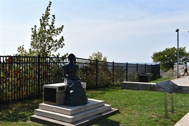 A bronze statue of a seated woman in a long dress is on a raised platform in a grassy park area. A black metal fence runs behind the statue, and a plaque is visible in the foreground.