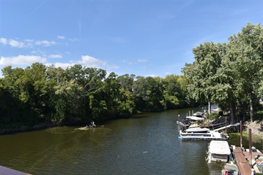 Boats docked on the right side of a wide, calm river with lush green trees lining the left bank under a bright blue sky.