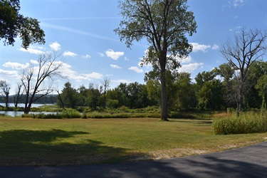 A wide grassy area with trees and a body of water in the background under a partly cloudy sky.