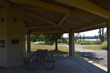 A covered picnic shelter with a table and benches, overlooking a lake and surrounding trees.