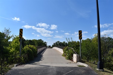 A paved pedestrian bridge arches over a path, flanked by lush green trees and bushes under a bright blue sky with scattered white clouds. Yellow and black warning signs are visible on either side of the bridge entrance.