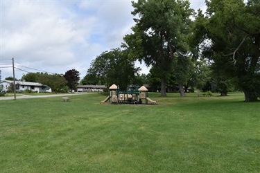 A children's playground with slides and climbing structures sits on a grassy lawn under a cloudy sky, with trees and houses in the background.