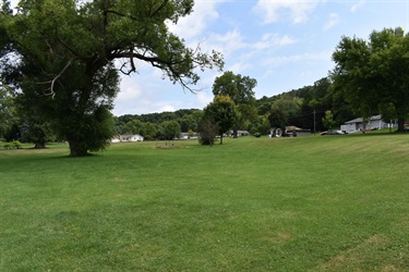 A wide, grassy lawn stretches towards a tree-lined hill with houses nestled among the trees under a partly cloudy sky.