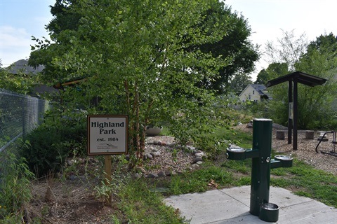A green drinking fountain for people and pets stands on a concrete pad. A sign reads 