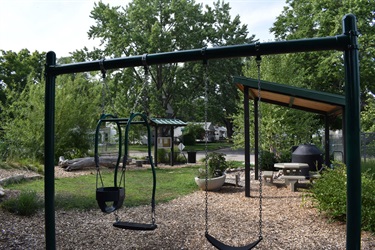 A park with swings, a picnic table, and a covered shelter surrounded by trees and greenery.