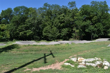 A dirt pump track with gentle curves and mounds is nestled among trees. The track is made of smooth, gray asphalt and is surrounded by green grass and rocks. A shadow of a pole is cast across the foreground.
