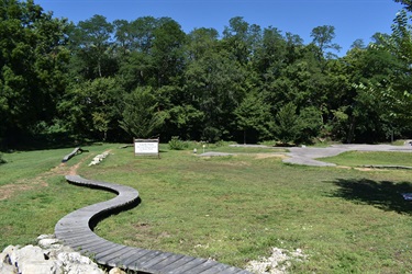 A wooden boardwalk curves through a grassy area with trees in the background and a sign in the distance.