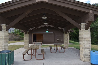A covered picnic area with wooden tables and benches, supported by stone pillars and a brown wooden roof structure.