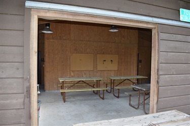 A covered outdoor picnic area with wooden picnic tables and bulletin boards on a wood-paneled wall.