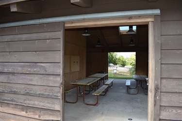 A covered picnic area with multiple wooden picnic tables and benches, looking out towards a grassy area with trees.