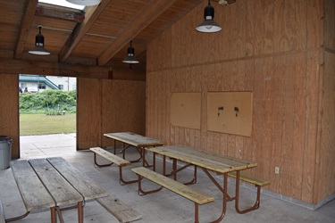 A covered outdoor picnic area with wooden picnic tables and benches, wood-paneled walls, and pendant lights. An open doorway reveals a grassy area and a building in the distance.