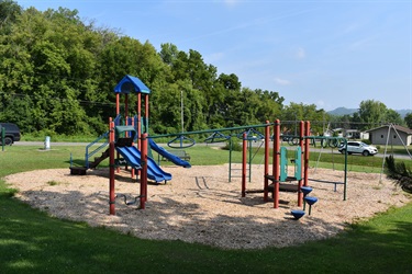 A bright blue and red playground set with slides, monkey bars, and swings, surrounded by wood chips and green grass, with trees and houses in the background.