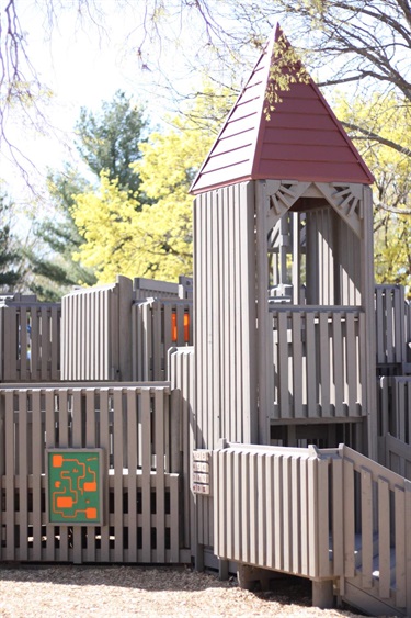 A wooden playground structure resembling a castle with a red roof and grey walls, featuring multiple levels and safety railings.