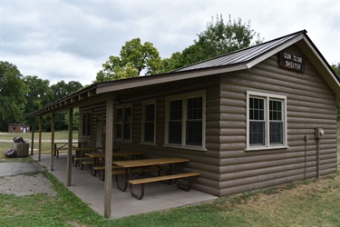 A small, brown, wood-sided building with a covered porch and picnic tables outside. The building has a metal roof and several windows. A sign on the building reads 