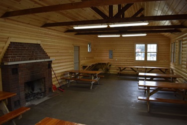 Interior of a wooden shelter building with picnic tables, a brick fireplace, and fluorescent lighting.