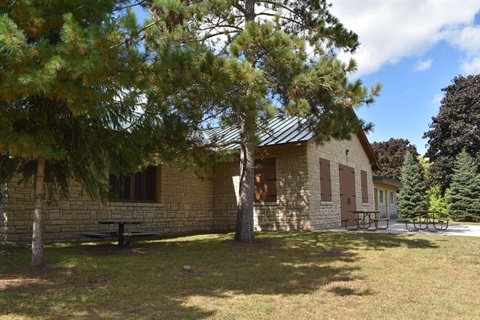 A stone building with a metal roof, surrounded by trees and picnic tables under a partly cloudy sky.