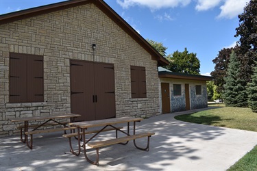 A stone building with large double doors and two windows is visible with picnic tables in front. A smaller section of the building with doors and windows is attached to the right.