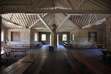 Interior of a large, open-air pavilion with picnic tables and a high, vaulted ceiling with exposed beams. A painting hangs on the far wall.