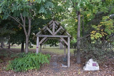 A wooden gazebo with an open structure and a bench inside, surrounded by trees and fallen leaves.