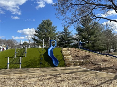 A modern playground with two slides, one blue and gray on a grassy hill and another blue slide on a smaller hill, with mulch and gravel ground cover.