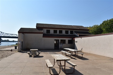 A building with picnic tables in front of it, a river and bridge in the background.