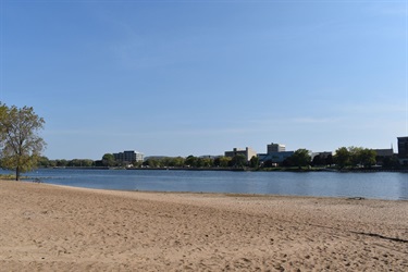 A wide sandy beach borders a calm blue river, with a distant treeline and several buildings visible under a clear sky.