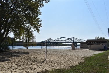 A sandy beach with volleyball nets in the foreground, a body of water and a bridge in the background, and a building on the right side of the image.