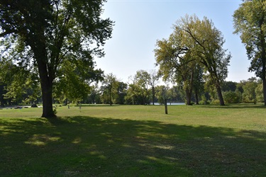 A wide grassy field with scattered trees and a body of water in the distance under a clear sky.