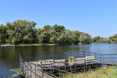 A wooden pier with benches sits on the edge of a calm river, surrounded by lush green trees under a clear blue sky.