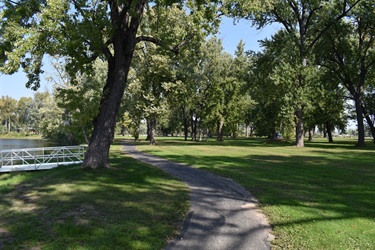 A paved walking path curves through a grassy park with mature trees and a body of water with a white dock on the left.