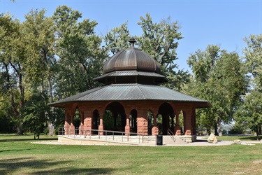 A circular stone pavilion with a domed roof sits in a grassy park surrounded by trees.