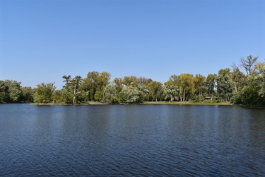 A wide, calm body of water reflects the clear blue sky and the lush green trees lining its distant shore.
