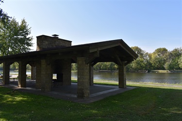 A covered picnic shelter with a stone fireplace and chimney next to a calm river on a sunny day.