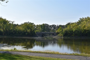 A calm body of water reflects the trees and a bridge in the distance under a clear sky.