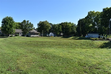 A wide, grassy lawn slopes upwards towards a line of houses and trees under a clear blue sky. A white garage with a dark roof is visible on the right side of the image.