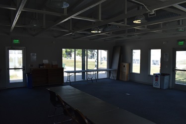 An empty multipurpose room with tables and chairs, large windows, and exit signs.