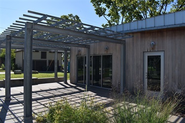 A modern building with a wooden exterior features a metal pergola extending over a paved patio area. Large windows and a glass door offer views into the interior.