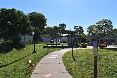 A paved pathway curves through a grassy park area with solar panel canopies and buildings in the background under a clear blue sky.