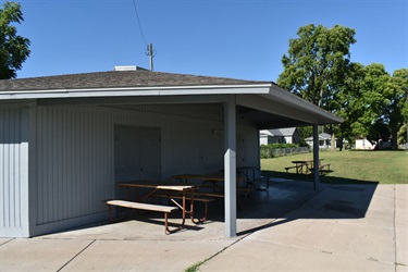 A covered picnic area with picnic tables and benches beside a building with a gray, slatted exterior. A grassy park with trees and houses is visible in the background under a clear blue sky.