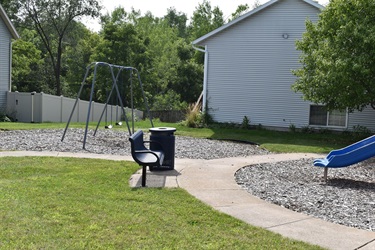 A playground with a swing set, a bench, a trash can, and a blue slide next to a building.